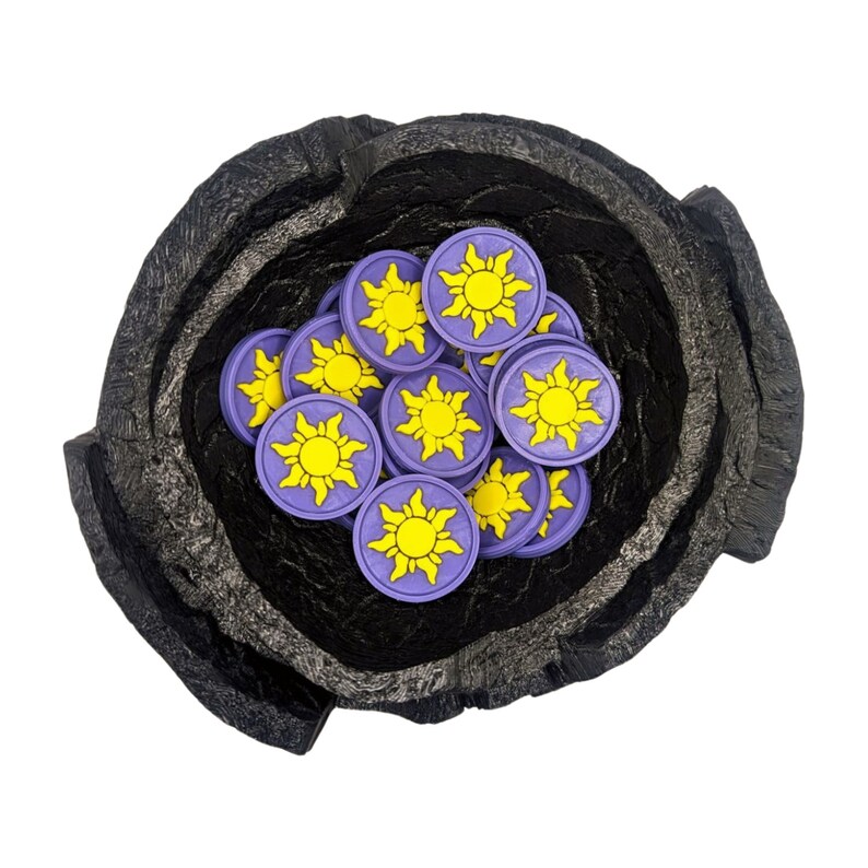 Puede incluir: Un cuenco negro texturizado lleno de fichas moradas y amarillas con forma de sol. Las fichas tienen un dise&ntilde;o de sol amarillo sobre un fondo morado. El cuenco tiene un aspecto rugoso y en capas.