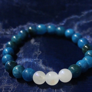 Puede incluir: Un brazalete de cuentas con piedras preciosas azules y blancas. El brazalete tiene una serie de cuentas azules con algunas cuentas azules más oscuras y tres cuentas blancas en el centro. El brazalete se muestra sobre un fondo azul oscuro.