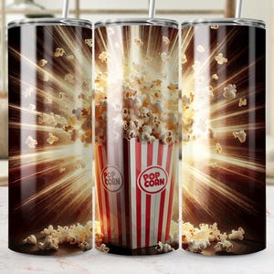 Puede incluir: Un vaso de acero inoxidable con un diseño de palomitas de maíz explotando de un cubo de palomitas de maíz a rayas rojas y blancas. El fondo es marrón oscuro con un brillo dorado.