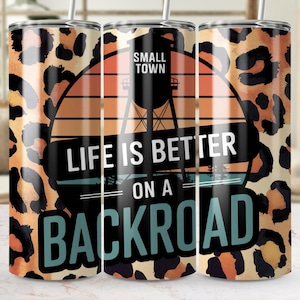 May include: Three stainless steel tumblers with a leopard print design and a sunset graphic. The tumblers have the text "Small Town" and "Life is Better on a Backroad".
