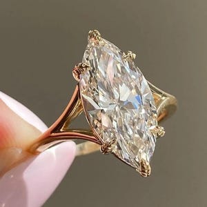 Puede incluir: Un anillo de oro con un gran diamante talla marquesa. El diamante es claro y multifacético, reflejando la luz. El anillo tiene un diseño delicado de caña dividida con pequeñas garras que aseguran la piedra.