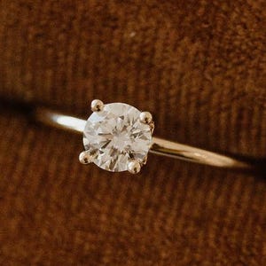 Może przedstawiać: Zbliżenie pierścionka z diamentem w oprawie solitaire. Pierścionek ma diament o okrągłym szlifie osadzony w czteroramiennej oprawie na cienkiej, polerowanej srebrnej obrączce. Diament błyszczy na brązowym tle.