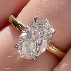 Könnte beinhalten: Ein goldener Ring mit einem großen, oval geschliffenen Diamanten. Der klare Diamant ist auf einem schlichten Goldband gefasst. Der Ring wird an einem Finger gezeigt, was das elegante Design hervorhebt.