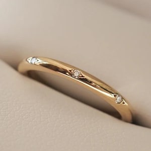 Puede incluir: Un anillo de oro con tres pequeñas piedras preciosas transparentes engastadas en la banda. El anillo se muestra dentro de un joyero beige claro. El anillo tiene un acabado liso y pulido y un diseño ligeramente curvado.