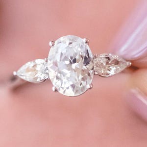 Könnte beinhalten: Ein funkelnder Verlobungsring mit einem großen, oval geschliffenen Diamanten in der Mitte, flankiert von zwei birnenförmigen Diamanten. Der Ring ist in einem silbernen Band gefasst, das die Brillanz der Diamanten zur Geltung bringt. Rosa Hintergrund.