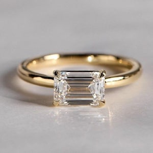 May include: A gold ring featuring a rectangular emerald-cut diamond. The ring has a polished gold band. The clear diamond reflects light, held in place by four prongs. The ring is presented on a light surface, showcasing its elegant design.