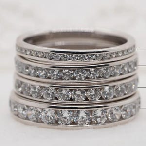 May include: A stack of four silver rings, each encrusted with small, clear gemstones. The rings vary in width, with text indicating sizes from 2.5mm to 3.2mm wide. The rings are displayed on a white surface.