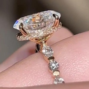 May include: A rose gold engagement ring with a large, oval-cut diamond. The diamond is encircled by a halo of smaller stones, with more diamonds set along the band. The ring's design is delicate and elegant, showcasing a high level of craftsmanship.