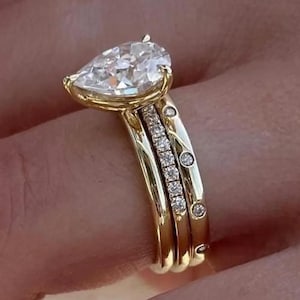 Puede incluir: Un conjunto de anillos de compromiso de oro con un diamante central en forma de pera. El anillo va acompañado de dos bandas de oro adicionales, una con pequeños diamantes y otra con tres pequeños diamantes. El anillo está en un dedo.