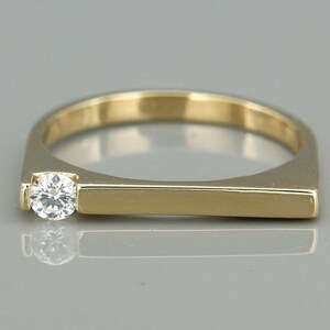 Może przedstawiać: Złoty pierścionek z pojedynczym, okrągłym, przezroczystym kamieniem szlachetnym osadzonym w środku obrączki.