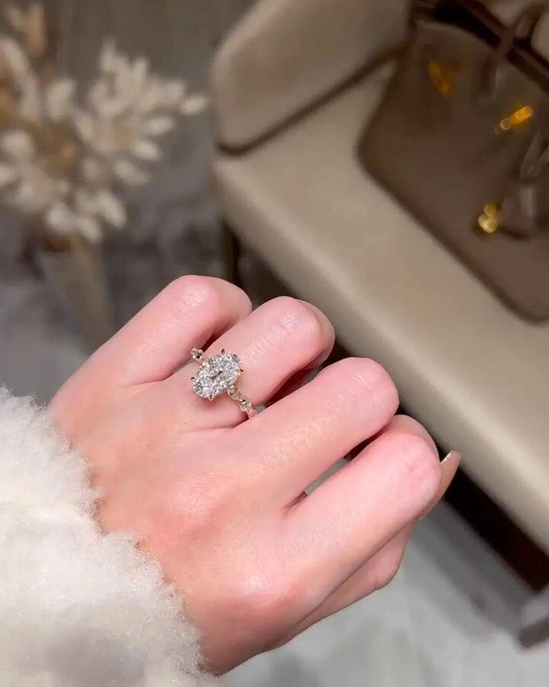 May include: A gold ring with an oval-cut diamond centre stone. The ring features a delicate band with smaller accent stones. The ring is worn on a finger, showcasing its elegant design and sparkle.