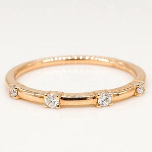 Puede incluir: Un anillo de banda de oro con cinco piedras preciosas redondas y transparentes. El anillo tiene una banda delgada y las piedras están engastadas en engastes cuadrados. El anillo se muestra sobre un fondo blanco.