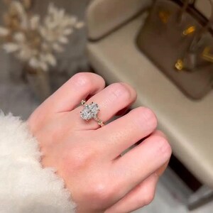 May include: A gold ring with an oval-cut diamond is shown on a hand. The ring's band features a delicate, twisted design. The diamond is the focal point, reflecting light. The background is blurred, emphasising the ring's elegance. The ring is a piece of jewellery.