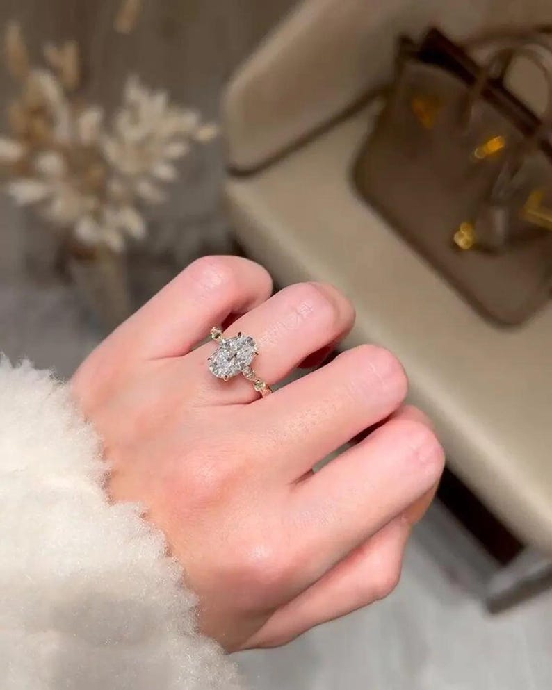 May include: A gold ring with an oval-cut diamond is shown on a hand. The ring's band features a delicate, twisted design. The diamond is the focal point, reflecting light. The background is blurred, emphasising the ring's elegance. The ring is a piece of jewellery.