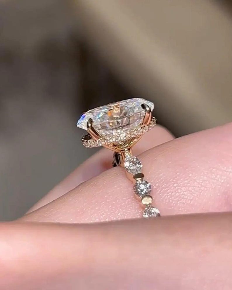 May include: A rose gold engagement ring with a large, oval-cut diamond. The diamond is encircled by a halo of smaller stones, with more diamonds set along the band. The ring's design is delicate and elegant, showcasing a high level of craftsmanship.