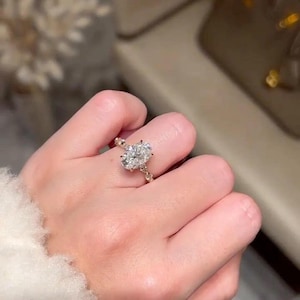May include: A gold ring featuring a large oval-cut diamond. The ring is set on a delicate band with smaller accent stones. The hand is wearing a white, fluffy sweater.