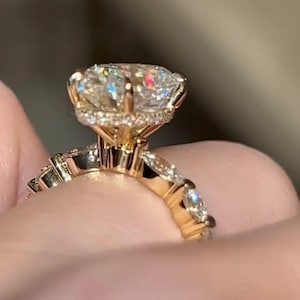 May include: A gold engagement ring with a large, round-cut diamond as the centerpiece. The ring features smaller diamonds set along the band and around the main stone, creating a sparkling effect. The ring is held up by a hand.