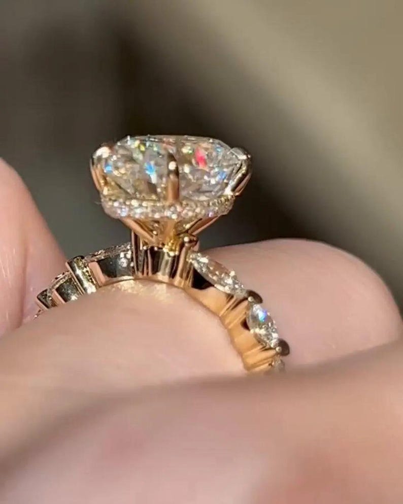 May include: A gold engagement ring with a large, round-cut diamond as the centerpiece. The ring features smaller diamonds set along the band and around the main stone, creating a sparkling effect. The ring is held up by a hand.