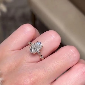 May include: A gold ring with an oval-cut diamond as the main stone. The ring's band has a detailed design with smaller accent stones. The diamond is held in place with prongs, and the ring is on a finger.