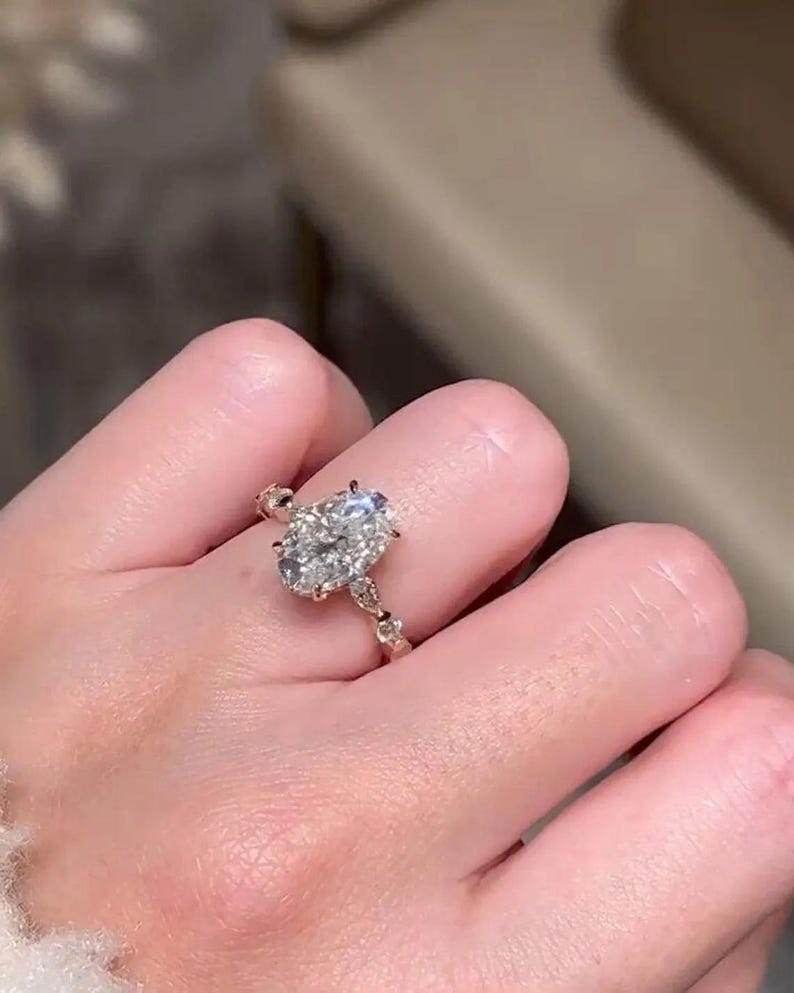 May include: A gold ring with an oval-cut diamond as the main stone. The ring's band has a detailed design with smaller accent stones. The diamond is held in place with prongs, and the ring is on a finger.