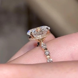 May include: A gold ring with a large, clear, oval-cut gemstone. The gemstone is surrounded by a halo of smaller stones. The ring's band is decorated with small stones, creating a sparkling effect. The ring is held by a hand.