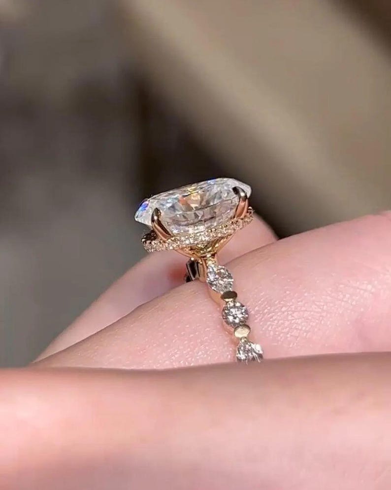 May include: A gold ring with a large, clear, oval-cut gemstone. The gemstone is surrounded by a halo of smaller stones. The ring's band is decorated with small stones, creating a sparkling effect. The ring is held by a hand.