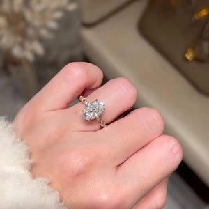 May include: A gold ring featuring an oval-cut diamond. The ring has smaller stones set into the band. The ring is on a finger, with a blurred background. The ring is a piece of jewellery.