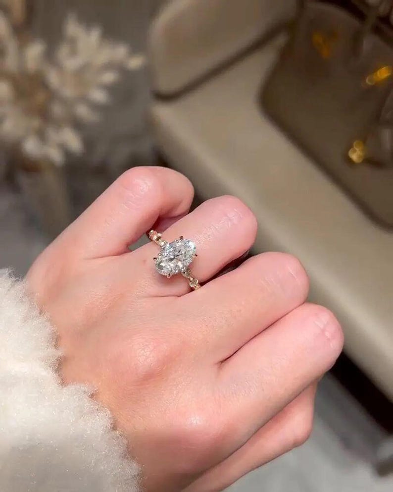 May include: A gold ring featuring an oval-cut diamond. The ring has smaller stones set into the band. The ring is on a finger, with a blurred background. The ring is a piece of jewellery.