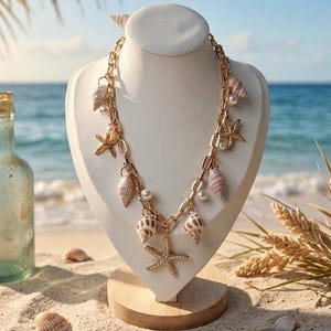 May include: A gold-toned necklace with seashell and starfish charms displayed on a white bust. The necklace features a chain design with various seashells, starfish, and pearl accents. The background shows a beach scene with sand, ocean, and a blue sky.