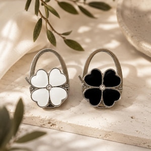 May include: Two foldable bag hooks with a silver-tone metal frame and a four-leaf clover design. One hook features white enamel petals, while the other features black enamel petals. Each clover center is accented with a small, clear, round crystal.