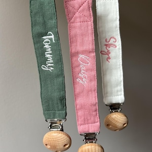 May include: Three personalised dummy clips in green, pink, and white. Each clip has a fabric strap with embroidered names: "Tommy", "Daisy", and "Skye". The clips feature a wooden bead and a metal clasp.