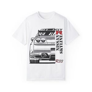 Puede incluir: Camiseta blanca con un gráfico en blanco y negro de un coche Nissan Skyline GT-R R33. El texto "GT-R", "NISSAN", "SKYLINE" y "R33" está impreso verticalmente en el lado derecho del coche.