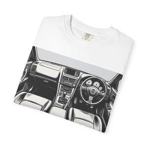 May include: White t-shirt with a monochrome illustration of a car interior, showcasing the dashboard, steering wheel, and seats. The graphic is centrally placed. A tag is visible at the neckline. The t-shirt is folded.
