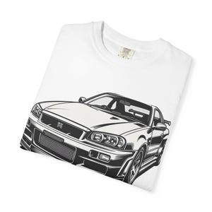 Puede incluir: Camiseta blanca con una ilustración en blanco y negro de un coche deportivo. El gráfico muestra la vista frontal y lateral del coche, destacando sus faros, parrilla y líneas de la carrocería. La camiseta está doblada.