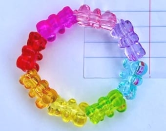 May include: A circular bracelet composed of translucent, multi-colored gummy bear-shaped beads. The beads are arranged in a rainbow sequence including red, orange, yellow, green, blue, purple, and pink, strung together to form a continuous loop.