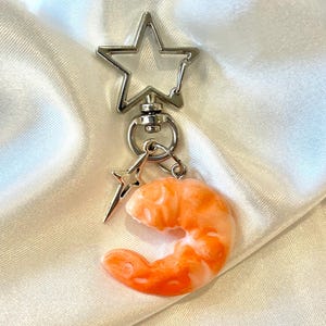 May include: A silver-toned metal keychain featuring a star-shaped clasp, a small four-pointed star charm, and a realistic orange and white shrimp-shaped pendant. The keychain components are connected by a circular ring and a swivel mechanism.