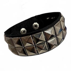 May include: A black wristband featuring two horizontal rows of metallic silver pyramid-shaped studs. The band is secured with two circular silver-toned metal snap fasteners visible on the interior side of the black material strap.