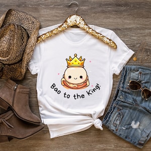 May include: White t-shirt featuring a cartoon bao bun wearing a gold crown, with the text "Bao to the King!". The design includes pink stars. The shirt is displayed with denim shorts, a brown hat, and boots.