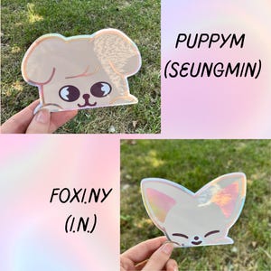 May include: Two holographic stickers featuring cartoon animal designs. One is a puppy with the text "PUPPYM (SEUNGMIN)". The other is a fox with the text "FOXI.NY (I.N.)". Both stickers have a pearlescent finish and are held up by a hand.