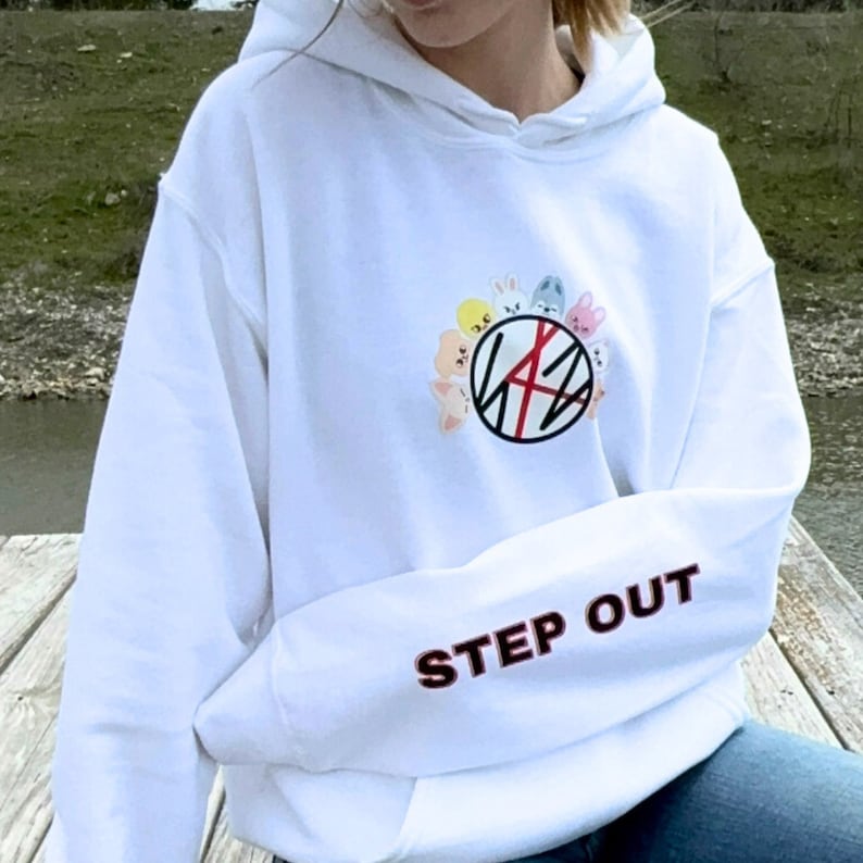 May include: White hooded sweatshirt with long sleeves. The left sleeve has the embroidered text "STEP OUT" in red and black. The front features a circular design with cartoon animal characters and a logo.