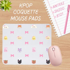 Könnte beinhalten: Ein rosa Mauspad mit hellblauem Hintergrund und einem sich wiederholenden Muster aus Comic-Tiergesichtern mit rosa Schleifen. Der Text "KPOP COQUETTE MOUSE PADS" befindet sich oben auf dem Mauspad. Eine rosa kabellose Maus befindet sich rechts neben dem Mauspad.