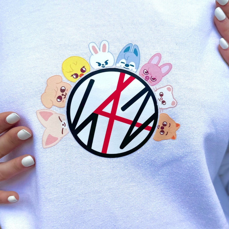 May include: White sweatshirt with a circular graphic featuring black and red lines and the letters "SKZ". The graphic is surrounded by cartoon animal characters in pink, blue, yellow, and brown.