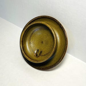 May include: A set of two ceramic dishes, a small bowl nested inside a larger plate. The dishes have a speckled olive green glaze with darker brown edges. The bowl and plate are round and appear handmade.