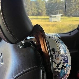 May include: A white helmet with various stickers and a brown leather strap attached to a black leather car seat.