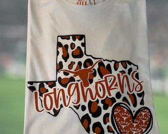 May include: White t-shirt featuring a Texas-shaped design with a leopard print pattern. The design includes the word "Longhorns" in a burnt orange script and a heart with the word "Texas" repeated. A tag is visible at the top.
