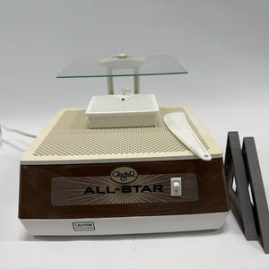May include: A glass grinder with a brown and beige base, a clear glass work surface, and a white plastic water reservoir. The text "ALL-STAR" is visible on the front. A white plastic spatula and two gray angle guides are also present.