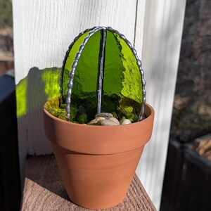 May include: A handcrafted stained glass cactus in a terracotta pot. The cactus features vibrant green glass panels framed with silver solder. The pot contains small stones and sits on a wooden surface, creating a decorative accent.