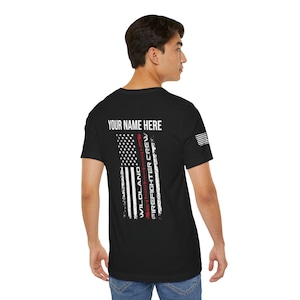 Puede incluir: Camiseta negra con un diseño de bandera estadounidense blanca y el texto "Wildland Firefighter Crew" impreso verticalmente en el lado derecho de la bandera. El texto "Your Name Here" está impreso sobre la bandera.