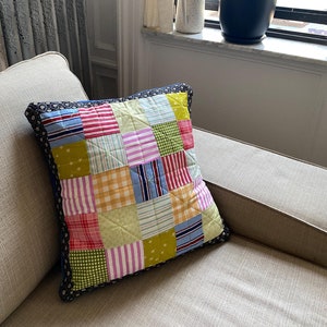 Może przedstawiać: Poduszka patchworkowa z różnymi kolorami i wzorami, w tym czerwonym, zielonym, żółtym, niebieskim i różowym. Poduszka ma ciemnoniebieski obrzeże i leży na jasnobrązowej kanapie.