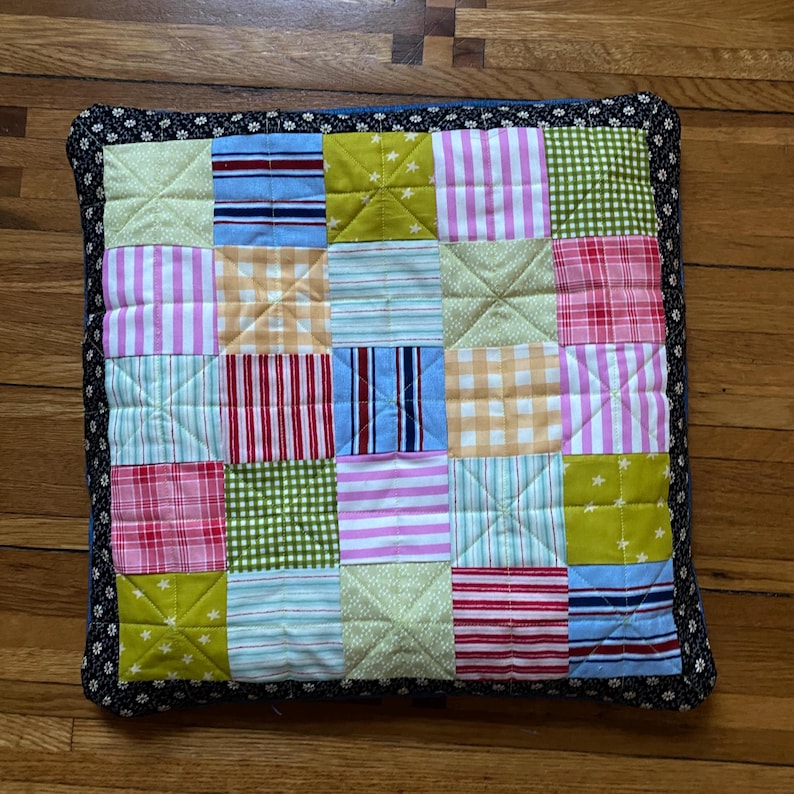 Pode incluir: Uma almofada de patchwork colorida com uma borda floral preta e branca. A almofada &eacute; feita de v&aacute;rios tecidos em tons de rosa, verde, azul, amarelo e vermelho. Os padr&otilde;es dos tecidos incluem listras, xadrez e bolinhas.